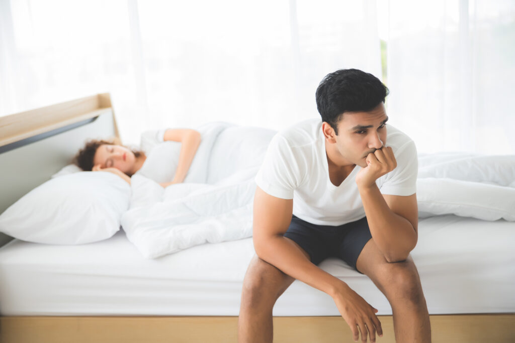 An Indian man was sitting. He had a strained facial expression and anxious. On the bed there was a sleeping woman.